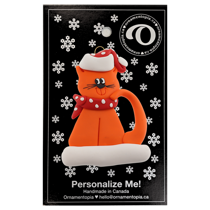 Ornament of orange cat in red and white polka dot Christmas hat and scarf, on display backing card.