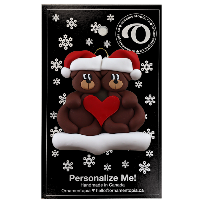 A couple’s ornament of two brown bears holding a red heart together, on display backing card.