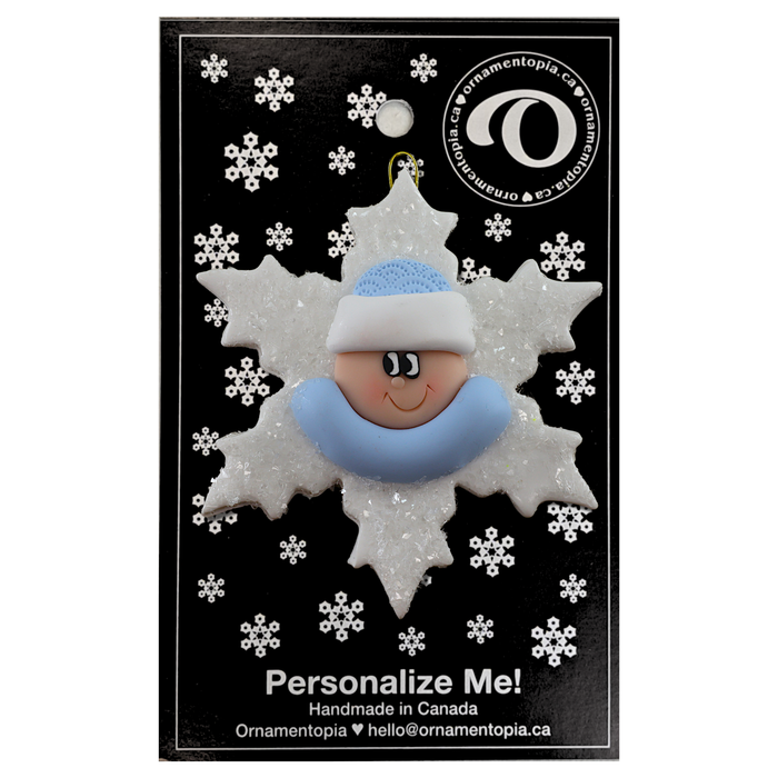 Baby ornament with infant face and blue hat on glittery white snowflake base, on display backing card.