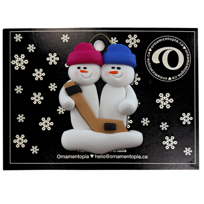 Family ornament with 2 white snowmen in bright hats, holding a hockey stick, on display backing card.
