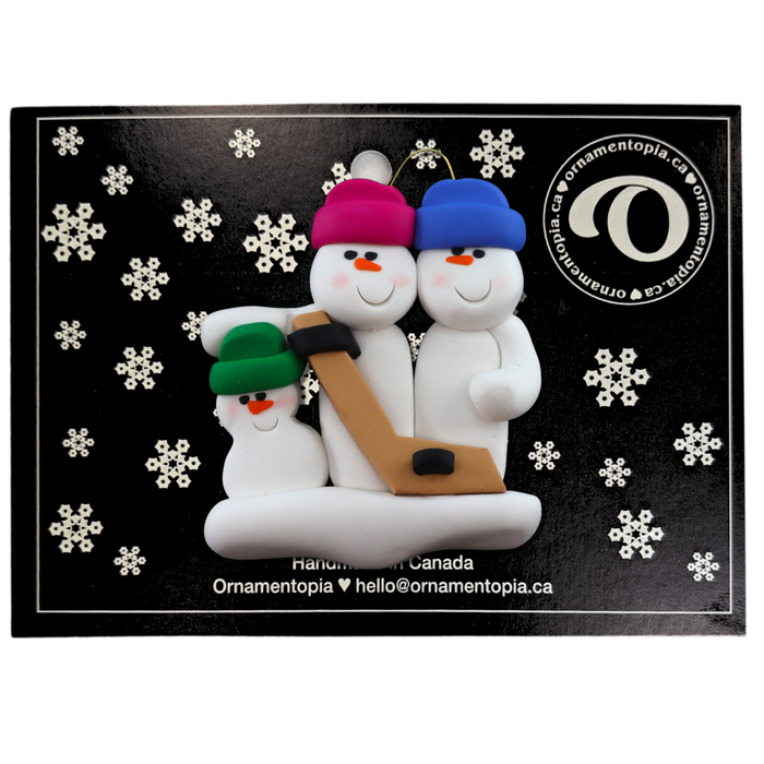 Family ornament with 3 white snowmen in bright hats, holding a hockey stick, on display backing card.