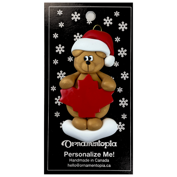 Tan bear in red festive hat with maple leaf ornament, on display backing card.