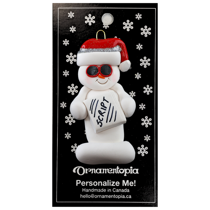 Ornament of white snowman in red hat and sunglasses holding a movie script, on display backing card.