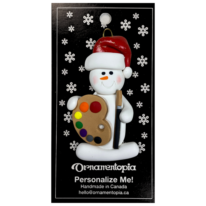 Ornament of white snowman in red hat holding a paint palette and paintbrush, on display backing card.