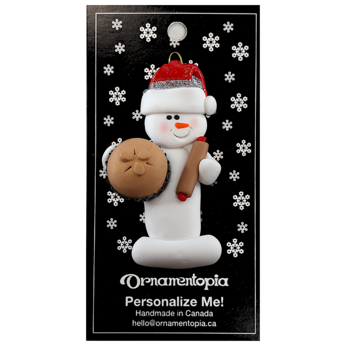 Ornament of white snowman in red hat holding a baked pie and rolling pin, on display backing card.