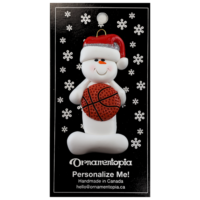 Ornament of white snowman in red hat holding a basketball, on display backing card.