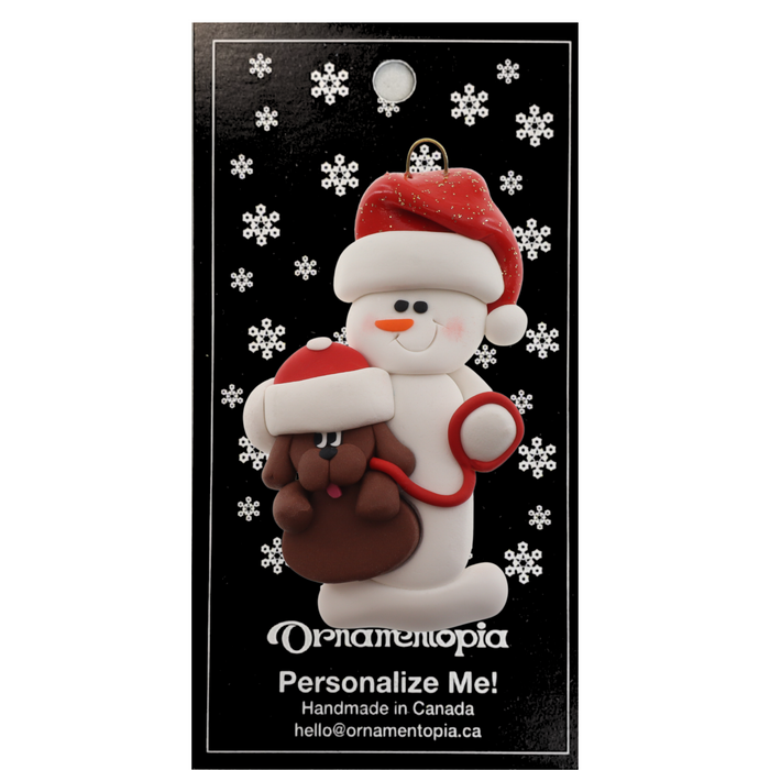 Ornament of white snowman in red hat holding a brown dog on a leash, on display backing card.