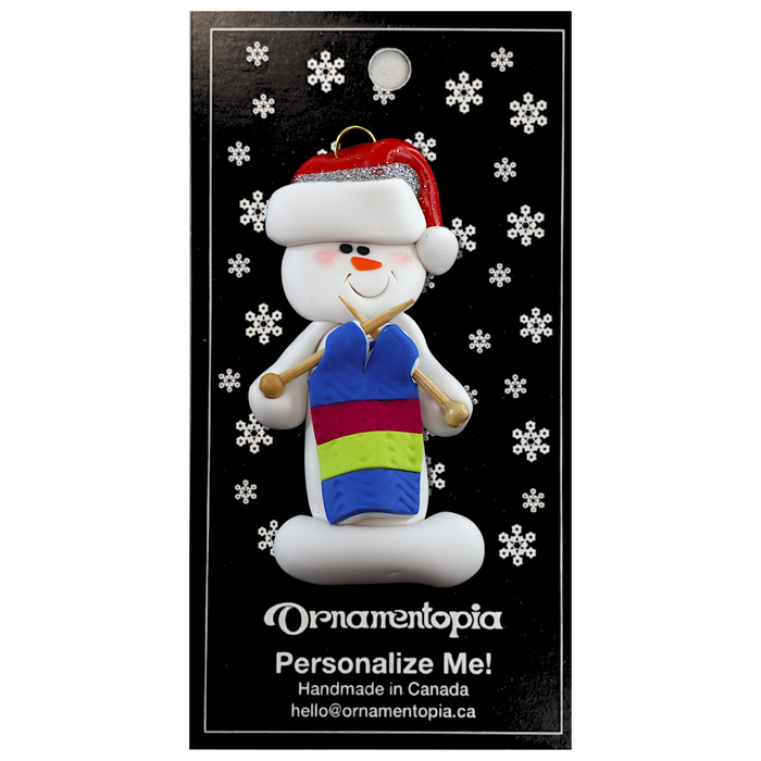 Ornament of white snowman in red hat holding an in-progress knitting project on knitting needles, on display backing card.