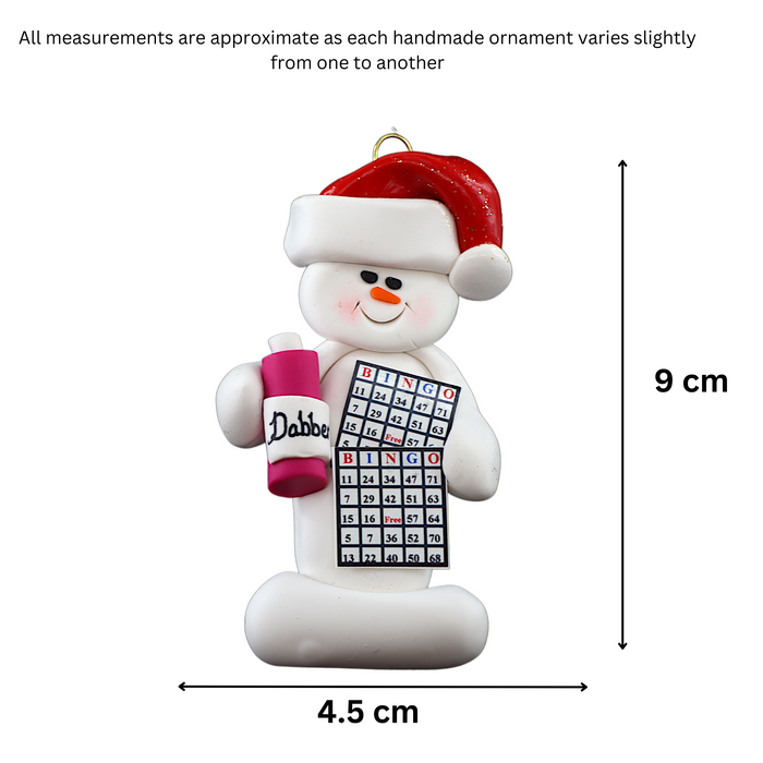 Ornament of white snowman in red hat holding a bingo card and dabber, graphed to show size (4.5x9cm).