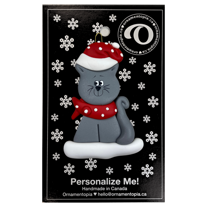 Ornament of grey cat in red and white polka dot Christmas hat and scarf, on display backing card.