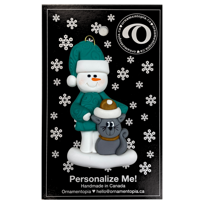 Ornament of snowman in a green shirt and hat with grey cat in Christmas hat, on display backing card.