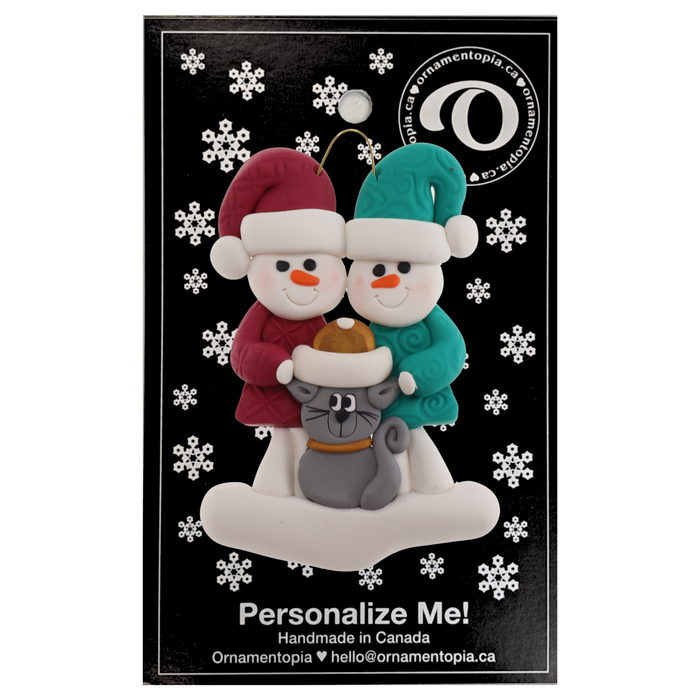 Ornament of snowman couple in red and green shirts and hats with grey cat in Christmas hat, on a display backing card.