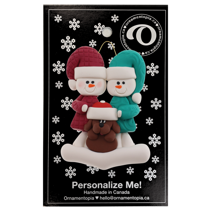 Ornament of snowman couple in red and green shirts and hats with brown dog in Christmas hat, on display backing card.