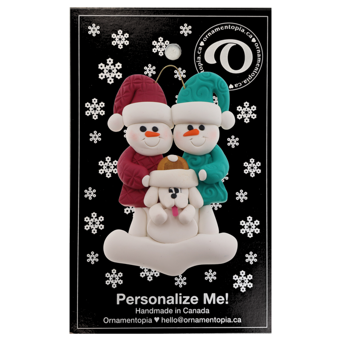 Ornament of snowman couple in red and green shirts and hats with white dog in Christmas hat, on display backing card.