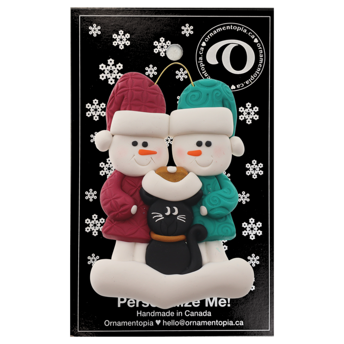 Ornament of snowman couple in red and green shirts and hats with a black cat in Christmas hat, on a display backing card.