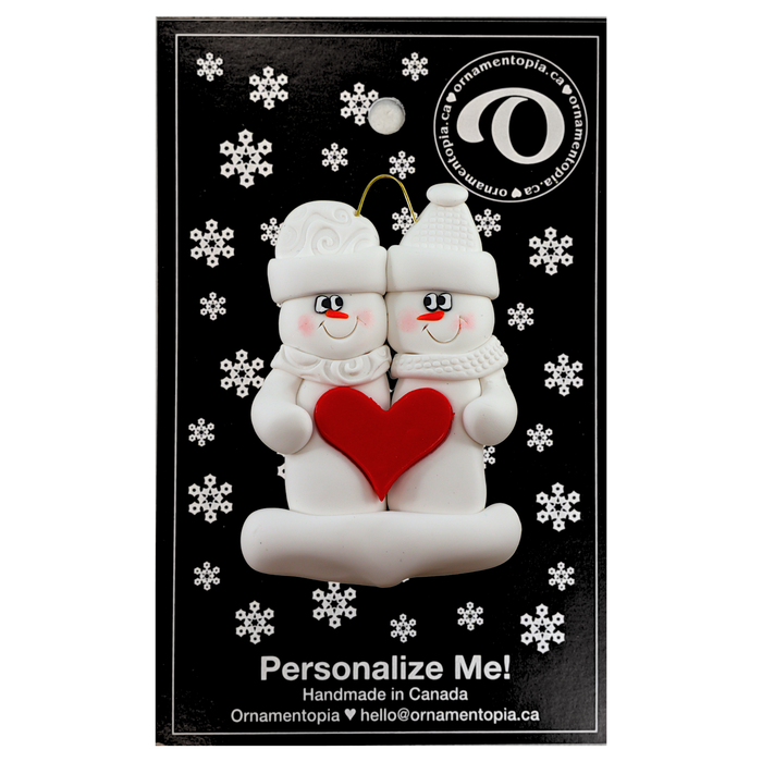 A couple’s ornament of two white snowmen holding a red heart together, on display backing card.