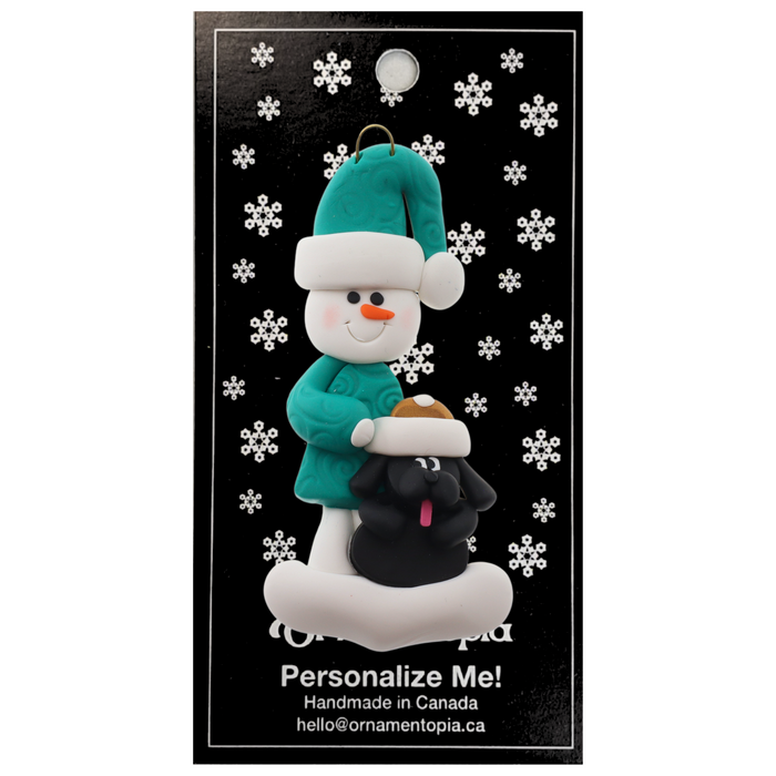 Ornament of snowman in green shirt and hat with black dog in Christmas hat, on display backing card.